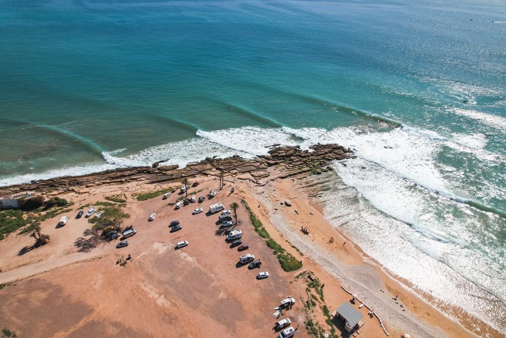 Waves breaking along a long beach, seen from above with a car park behind the sand