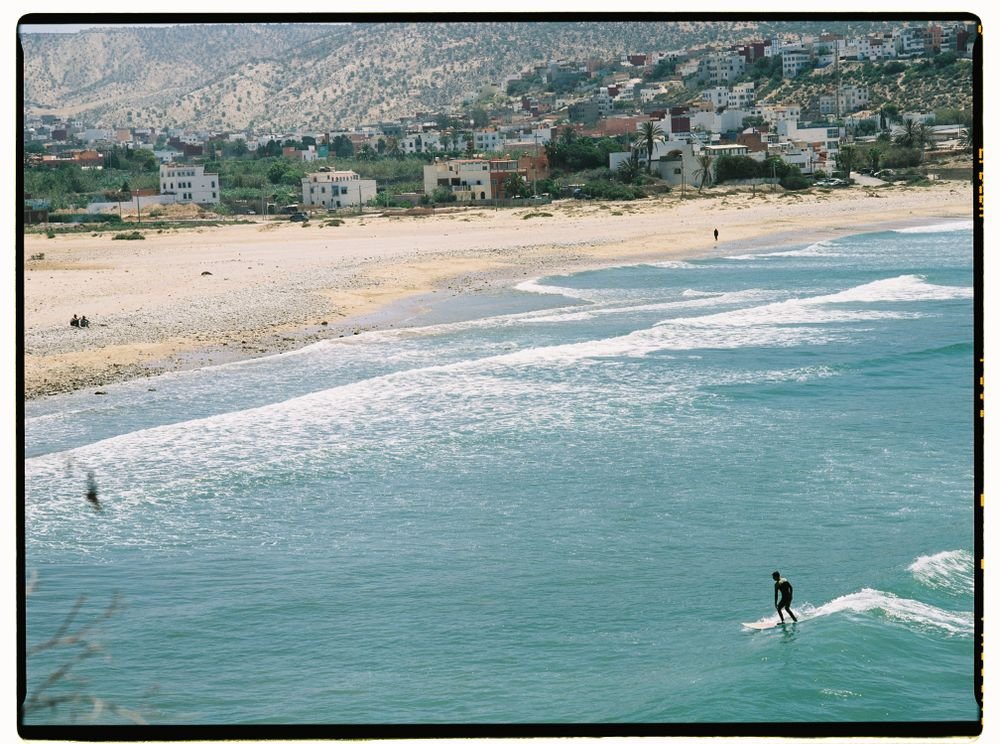 A surfer riding a wave over turquoise water with the beach and town behind