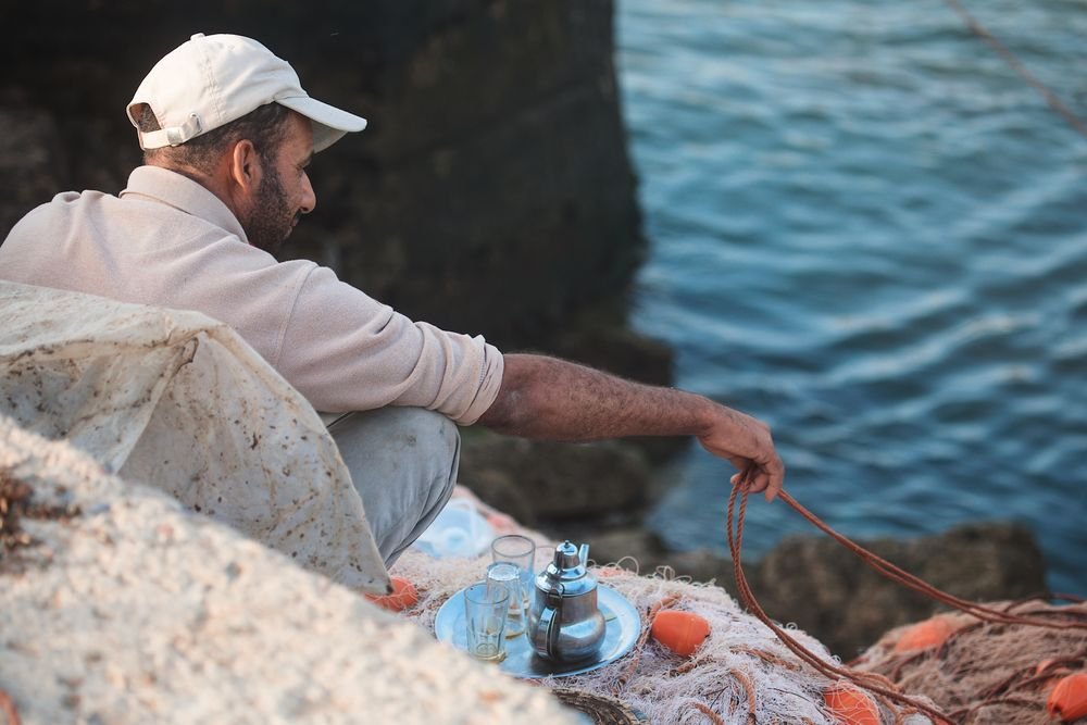 Un pescador remienda redes junto al agua, con una bandeja de té al lado
