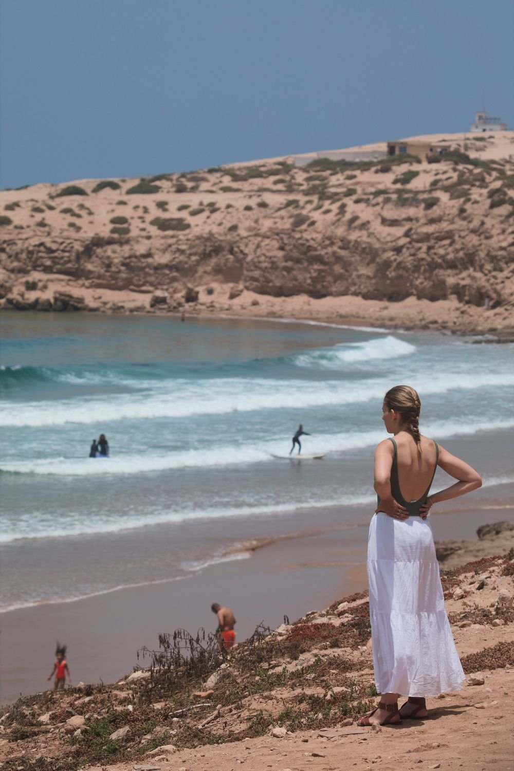 A woman in a white dress watching surfers from the rocks above the beach