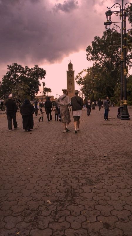 Pedestrians on a paved square in Marrakech with the Koutoubia minaret in the distance