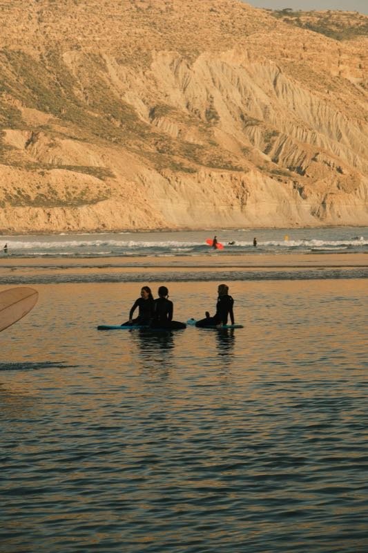 Surfers sitting on their boards in calm water below sandy cliffs