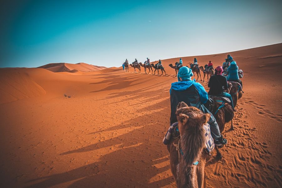 Camel train with riders in blue robes crossing sand dunes toward sunset