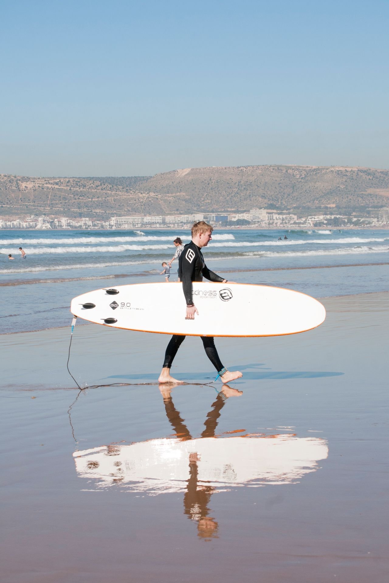 A surfer with a soft-top board on Agadir beach, the city skyline behind