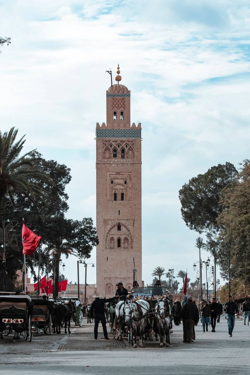 Kutubiyya Mosque minaret in Marrakesh with Moroccan red flags and horse-drawn carriages in the square below