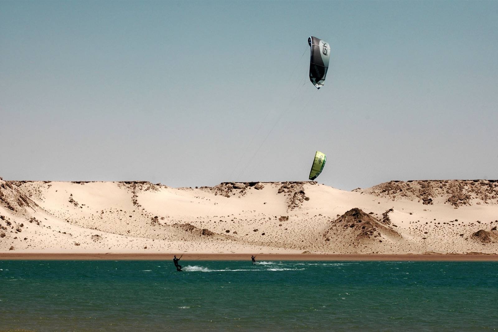 Two kite surfers on Dakhla's flat turquoise lagoon with desert sand dunes behind, Morocco